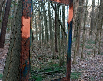 May include: A rectangular mirror with a copper frame, showing a reflection of a forest. The frame has a blue and green patina, and the mirror is hanging on a tree trunk.