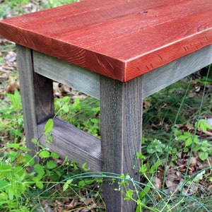 May include: A wooden bench with a red stained top and gray stained legs. The bench is sitting on a bed of green grass and brown leaves.