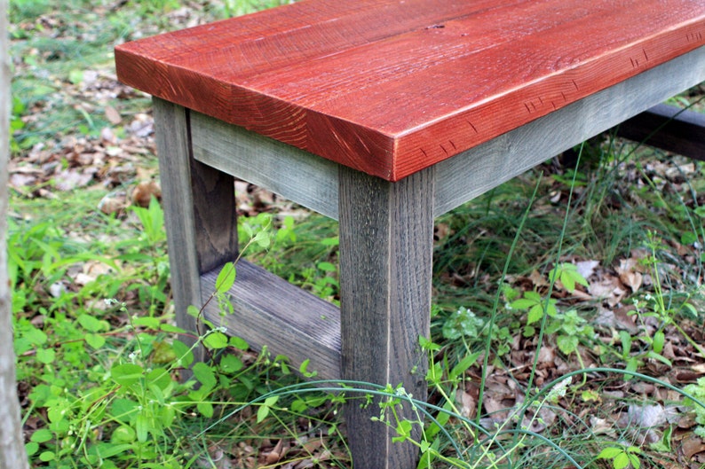 May include: A wooden bench with a red stained top and gray stained legs. The bench is sitting on a bed of green grass and brown leaves.