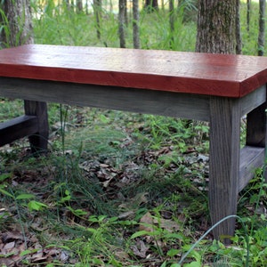 May include: A rustic wooden bench with a red stained top and gray stained legs. The bench is sitting on a bed of green grass and leaves.