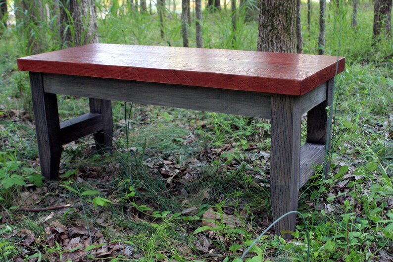 May include: A rustic wooden bench with a red stained top and gray stained legs. The bench is sitting on a bed of green grass and leaves.