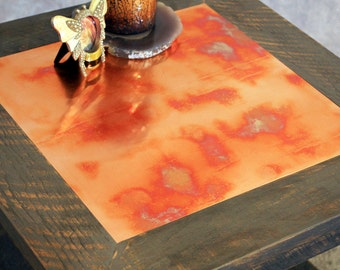 May include: A square wooden table with a copper top. The copper has a reddish-orange patina and is framed by a dark wood border.