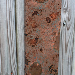 May include: A close-up of a rectangular piece of copper metal with a patina finish. The copper has a mottled appearance with shades of brown, orange, and green.