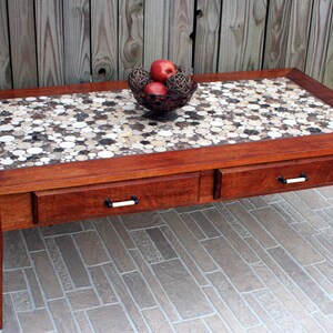May include: A wooden coffee table with two drawers and a mosaic tile top. The tile is made of small, round pieces of wood in shades of brown and white. A bowl of red apples and decorative balls sits on the table.