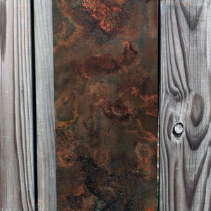 May include: A rusty metal sheet with a mottled brown, orange, and green patina. The sheet is mounted on a wooden fence.