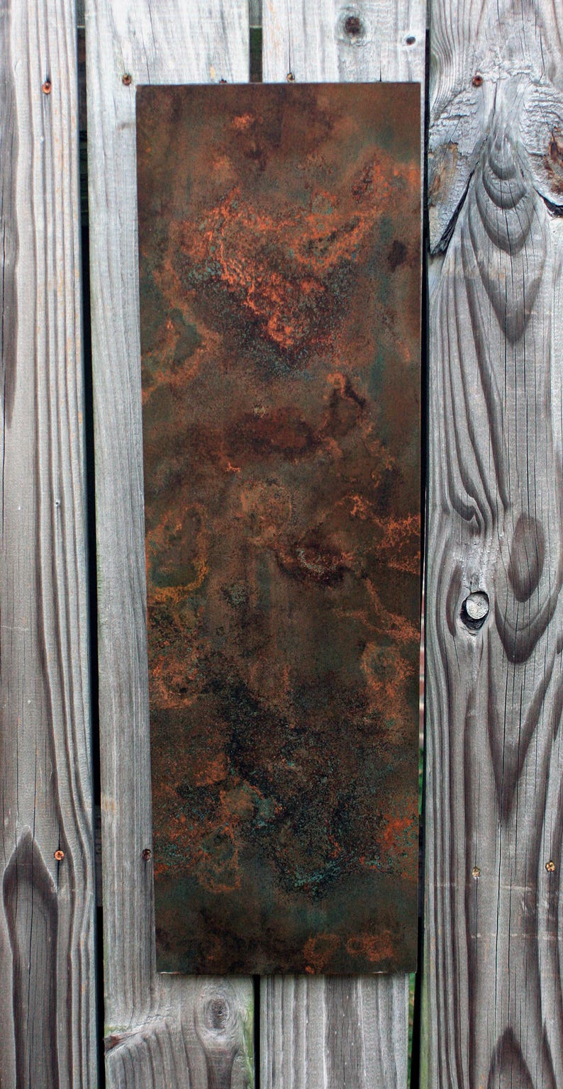 May include: A rusty metal sheet with a mottled brown, orange, and green patina. The sheet is mounted on a wooden fence.