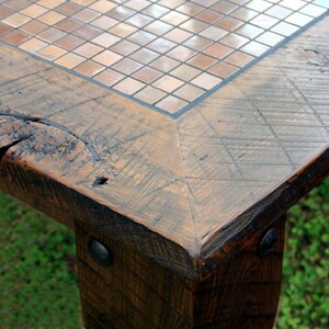 May include: Close-up of a wooden table corner with a mosaic tile top. The table has a rustic, distressed look with visible wood grain and knots. The tile top is made of small, square tiles in shades of brown and beige.