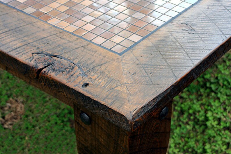 May include: Close-up of a wooden table corner with a mosaic tile top. The table has a rustic, distressed look with visible wood grain and knots. The tile top is made of small, square tiles in shades of brown and beige.