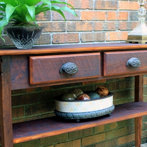 May include: A rustic wooden console table with two drawers and a lower shelf. The table has a dark brown stain and features black metal drawer pulls. The table is sitting in front of a brick wall.