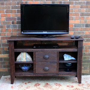 May include: A dark brown wooden TV stand with two drawers and open shelves. The stand has a flat top surface with a black flat screen TV on top. The TV stand is in front of a brick wall.