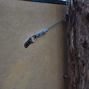 May include: A close-up of a brown wooden board with a metal hook and blue string attached to it. The hook is attached to a tree trunk.