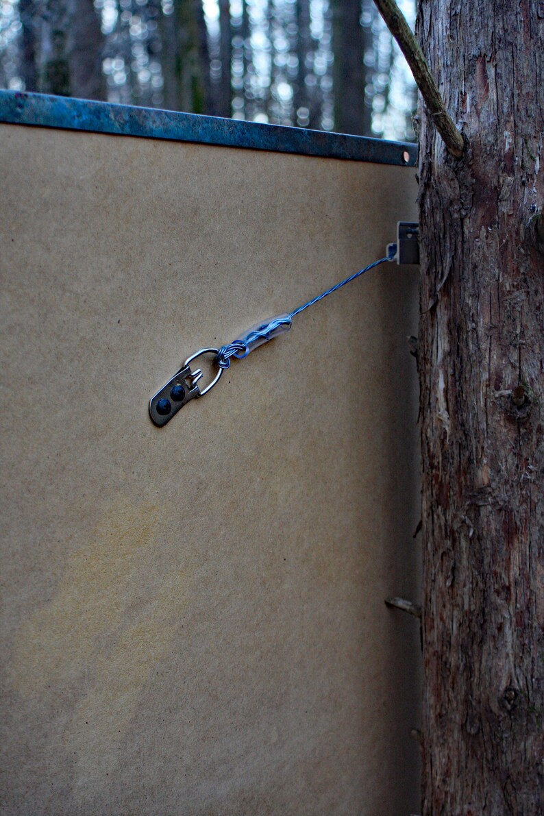May include: A close-up of a brown wooden board with a metal hook and blue string attached to it. The hook is attached to a tree trunk.