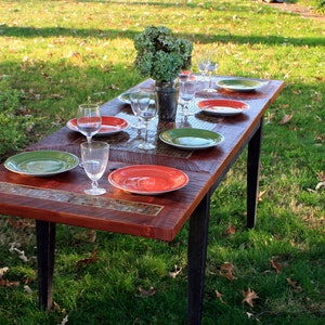K&ouml;nnte beinhalten: Ein rustikaler Holztisch mit schwarzen Metallbeinen ist f&uuml;r ein Essen im Freien gedeckt. Der Tisch ist mit einem Mosaik-Fliesendesign verziert und verf&uuml;gt &uuml;ber gr&uuml;ne und rote Teller, Gl&auml;ser und eine Vase mit gr&uuml;nen Pflanzen.