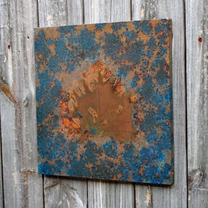 May include: A square metal wall art piece with a blue and brown rust-like pattern. The center of the piece has a large, brown, organic shape.