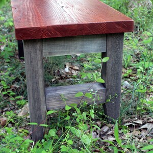 May include: A rustic wooden bench with a red stained top and gray stained legs. The bench is sitting on a bed of green grass and brown leaves.