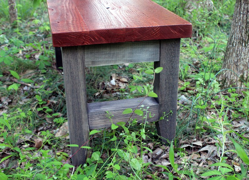 May include: A rustic wooden bench with a red stained top and gray stained legs. The bench is sitting on a bed of green grass and brown leaves.