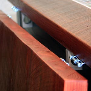 K&ouml;nnte beinhalten: Ein Holztisch mit einer Klappverl&auml;ngerung. Die Tischplatte ist aus dunklem Holz mit einem r&ouml;tlichen Farbton. Die Klappverl&auml;ngerung ist mit Metallscharnieren am Tisch befestigt.