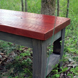 May include: A wooden bench with a red stained top and gray stained legs. The bench is in a grassy area with trees in the background.