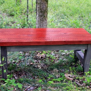 May include: A rustic wooden bench with a red stained top and gray stained legs. The bench is sitting on a bed of green grass and leaves.
