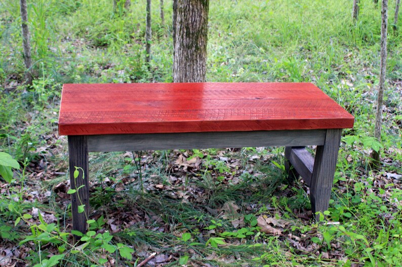 May include: A rustic wooden bench with a red stained top and gray stained legs. The bench is sitting on a bed of green grass and leaves.