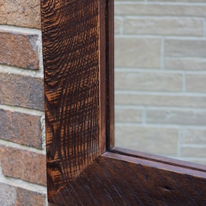 May include: Close-up of a dark brown wooden frame with a mirror inside. The frame is made of reclaimed wood with a rustic finish. The frame is mounted on a brick wall.