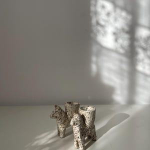 Peut inclure: Deux bougeoirs en céramique en forme d'animaux avec une finition tachetée crème et brune. Chaque pièce présente un design de créature à quatre pattes avec une ouverture cylindrique sur le dos pour une bougie, avec une surface texturée.
