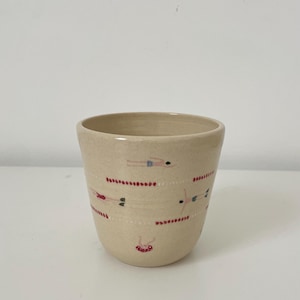 Op de afbeelding: Een beige keramieken beker met handgeschilderde illustraties van kleine figuren die in verschillende posities zwemmen. Het ontwerp bevat horizontale stippellijnen in rood en wit, afgewisseld met zwemmers in kleurrijke zwemkleding.