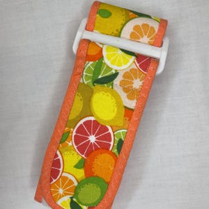 May include: A fabric wrist strap featuring a vibrant citrus fruit pattern with oranges, lemons, grapefruits, and limes. The strap has an orange border and a white plastic buckle. The strap is approximately 10 inches long.