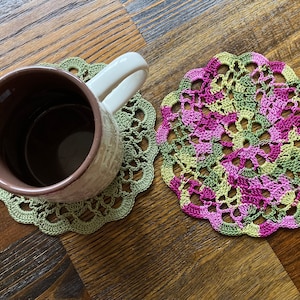 May include: A brown ceramic mug with a white handle rests on a green crocheted coaster. A second coaster, in shades of pink, green, and yellow, sits beside it. Both coasters have a scalloped edge and a detailed, openwork design.