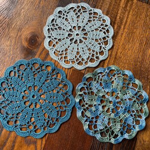 May include: Three crocheted doilies in shades of blue and green. The doilies are circular and have a delicate lace pattern.