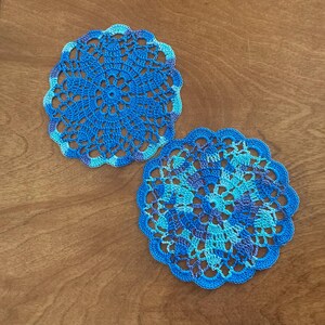 May include: Two handmade, blue crochet doilies with intricate floral patterns. The doilies feature shades of blue and purple, with scalloped edges. They are placed on a wooden surface.