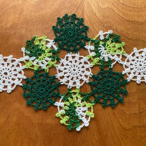 May include: A white and green crocheted doily with a floral design. The doily is made up of several smaller circles that are connected together.