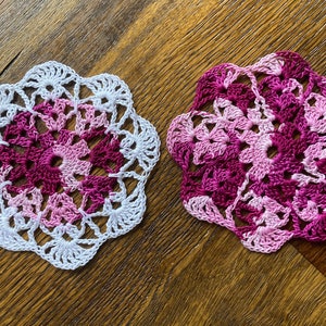 May include: Two crocheted doilies, one white with a pink and purple center, and one pink and purple with a white border.