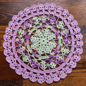 May include: A handmade, circular doily in shades of lavender and light green. The crochet design features a central floral pattern, surrounded by intricate openwork and a scalloped edge. The doily is approximately 12 inches in diameter.
