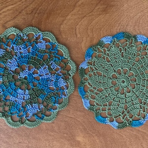 May include: Two handmade crochet doilies. One is primarily green with blue and green accents, the other is green with blue accents. Both have a scalloped edge and intricate openwork patterns. The doilies are displayed on a wooden surface.