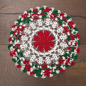 May include: A round, hand-crocheted doily featuring a central red floral motif surrounded by concentric rings of white and green yarn. The intricate lace pattern radiates outward, ending in a scalloped edge with alternating red, white, and green.