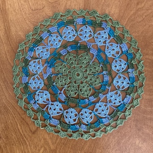 May include: A handmade, circular crochet doily in shades of green, blue, and purple. The doily features a floral pattern with a scalloped edge. The color palette includes olive green, teal, and periwinkle, creating a decorative textile.