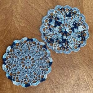 May include: Two handmade, round crochet doilies. The doilies are light blue with navy blue accents and a scalloped edge. The intricate openwork design is visible. The doilies are placed on a wooden surface.