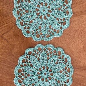May include: Two handmade, round crochet doilies in a light teal color. The doilies feature an intricate openwork design with scalloped edges. They are displayed on a wooden surface, showcasing the detailed craftsmanship of the textile.