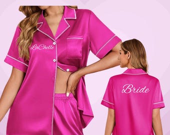 May include: A hot pink satin pajama set with white trim. The set includes a short-sleeved button-up shirt and matching shorts. The shirt has "La Chelle" embroidered on the front. The back of the shirt says "Bride". Text at the top says "Satin Pajama Set".