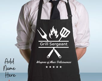 May include: Black apron with white text and graphics. The apron features the words "Grill Sergeant" and "Weapons of Mass Deliciousness." The text "4 Designs Available" is at the top. The name "Parker" is at the bottom.