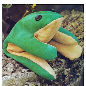 May include: A green and brown frog puppet with black button eyes. The puppet is sitting on a tree trunk.