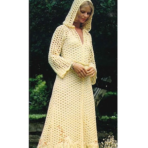 Pode incluir: Um vestido longo de crochê amarelo com capuz. O vestido tem um caimento solto e uma saia fluida. A modelo está em pé num jardim com folhagem verde e flores brancas.