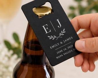 May include: A rectangular black metal bottle opener card featuring a white printed design with the initials E and J, a leaf motif, and the text EMILY & JAMES, JUNE 14, 2025, NAPA VALLEY, CA. The opener is positioned over a brown glass bottle cap.
