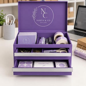 May include: A purple metal desk organizer with two drawers and an open top compartment. The lid features a white logo with the text NORTH & CO. EST. 2024. The organizer contains stationery items including cards, pens, tape rolls, and scissors.