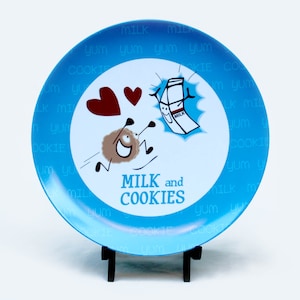 Puede incluir: Un plato de cerámica azul con una ilustración de dibujos animados de una galleta y una caja de leche. El plato tiene las palabras "MILK and COOKIES" escritas en él. El plato está en un soporte negro.