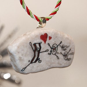 K&ouml;nnte beinhalten: Ein wei&szlig;er Stein-Anh&auml;nger mit einem handgemalten Design eines l&auml;chelnden Speckst&uuml;cks mit zwei Herzen dar&uuml;ber und drei l&auml;chelnden Eierfiguren, die davonlaufen. Der Anh&auml;nger h&auml;ngt an einer rot-wei&szlig;-gr&uuml;nen Schnur.