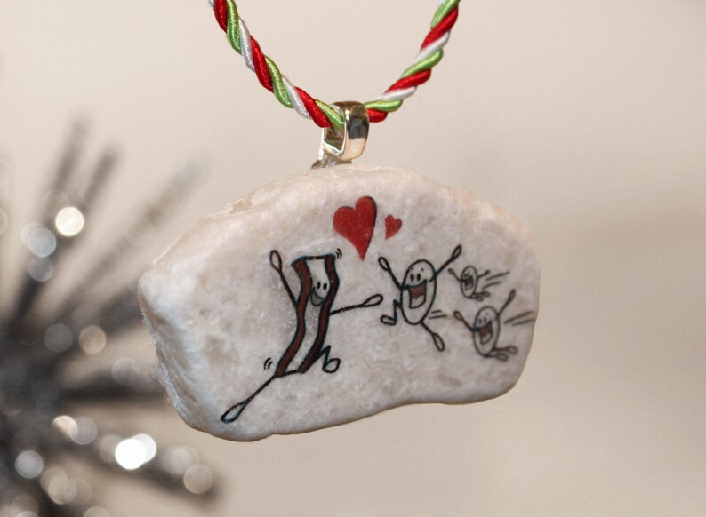 K&ouml;nnte beinhalten: Ein wei&szlig;er Stein-Anh&auml;nger mit einem handgemalten Design eines l&auml;chelnden Speckst&uuml;cks mit zwei Herzen dar&uuml;ber und drei l&auml;chelnden Eierfiguren, die davonlaufen. Der Anh&auml;nger h&auml;ngt an einer rot-wei&szlig;-gr&uuml;nen Schnur.