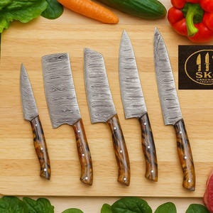 Könnte beinhalten: Ein Set aus fünf Küchenmessern aus Damaststahl mit Holzgriffen, angeordnet auf einem Holzschneidebrett. Die Klingen haben ein ausgeprägtes Muster. Die Griffe sind warmbraun. Das Bild enthält frisches Gemüse und das Logo SKC Handmade Creative.