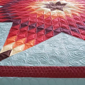 Puede incluir: Un edredón grande y colorido con un diseño de estrella. La estrella está compuesta por parches en forma de diamante en tonos de rojo, naranja y marrón, sobre un fondo azul claro con un patrón de remolino. El borde es de color rojo oscuro.